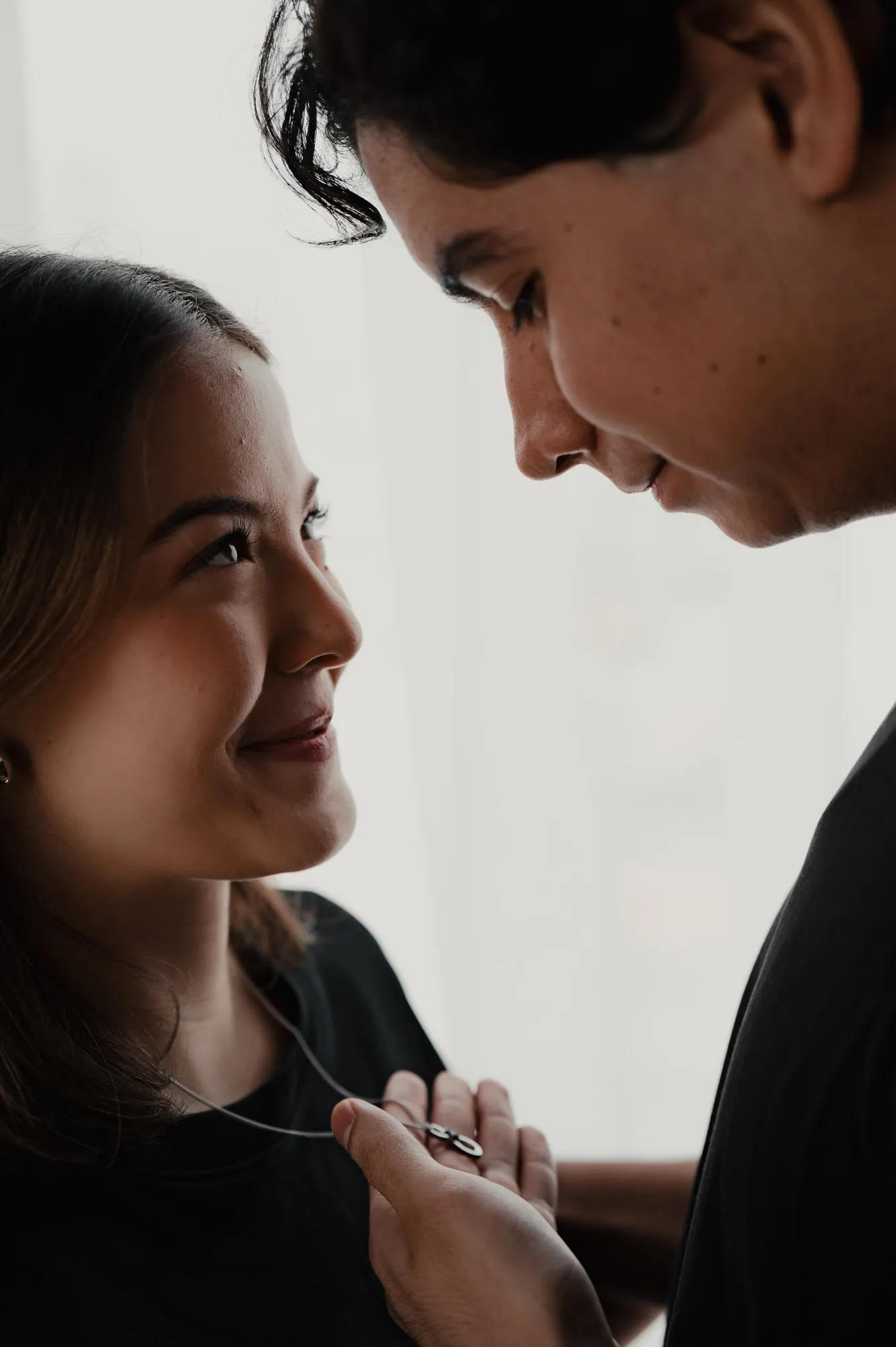 Primer plano de una pareja que se sonríe, él con la cabeza inclinada, junto a una ventana.