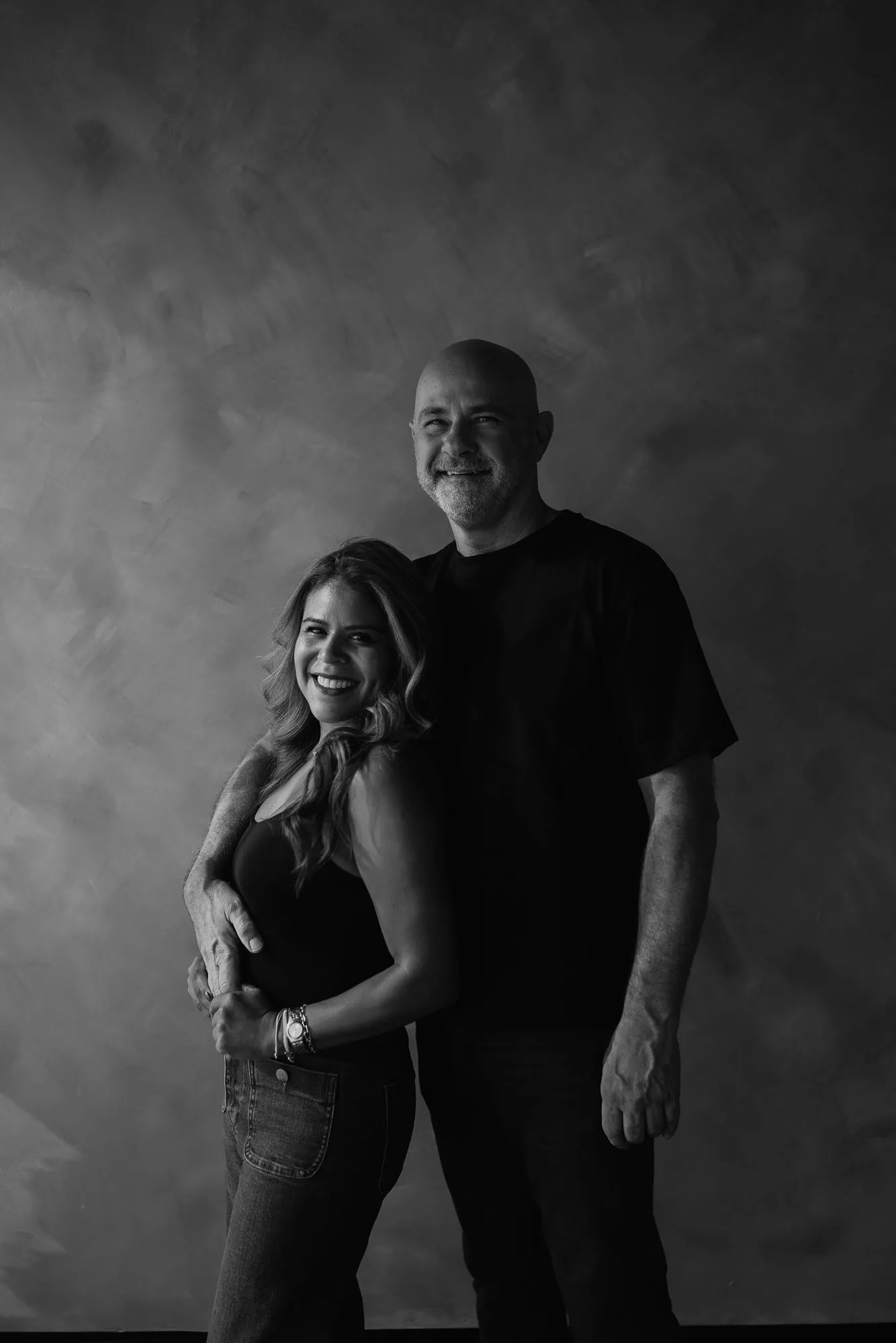 En blanco y negro, un hombre y una mujer posan abrazados frente a una pared texturizada oscura.