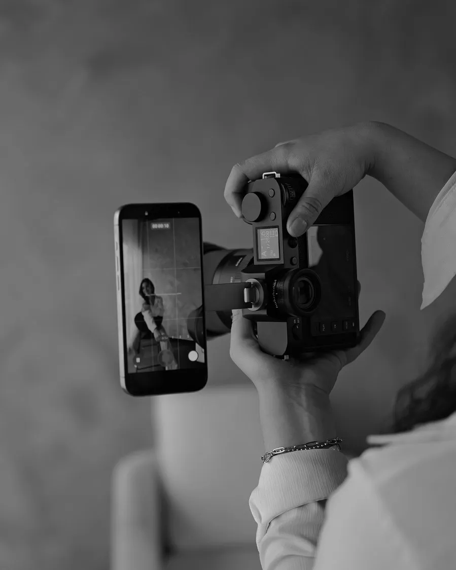 En blanco y negro, una mujer sentada en una silla sonríe a la cámara mientras una fotógrafa la graba con un teléfono montado sobre su cámara.
