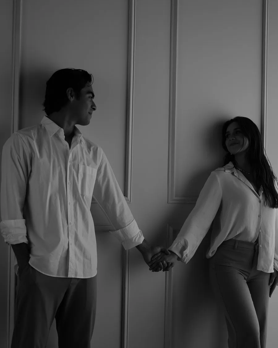 En blanco y negro, una pareja se toma de la mano, recargada en una pared con molduras junto a una cortina.