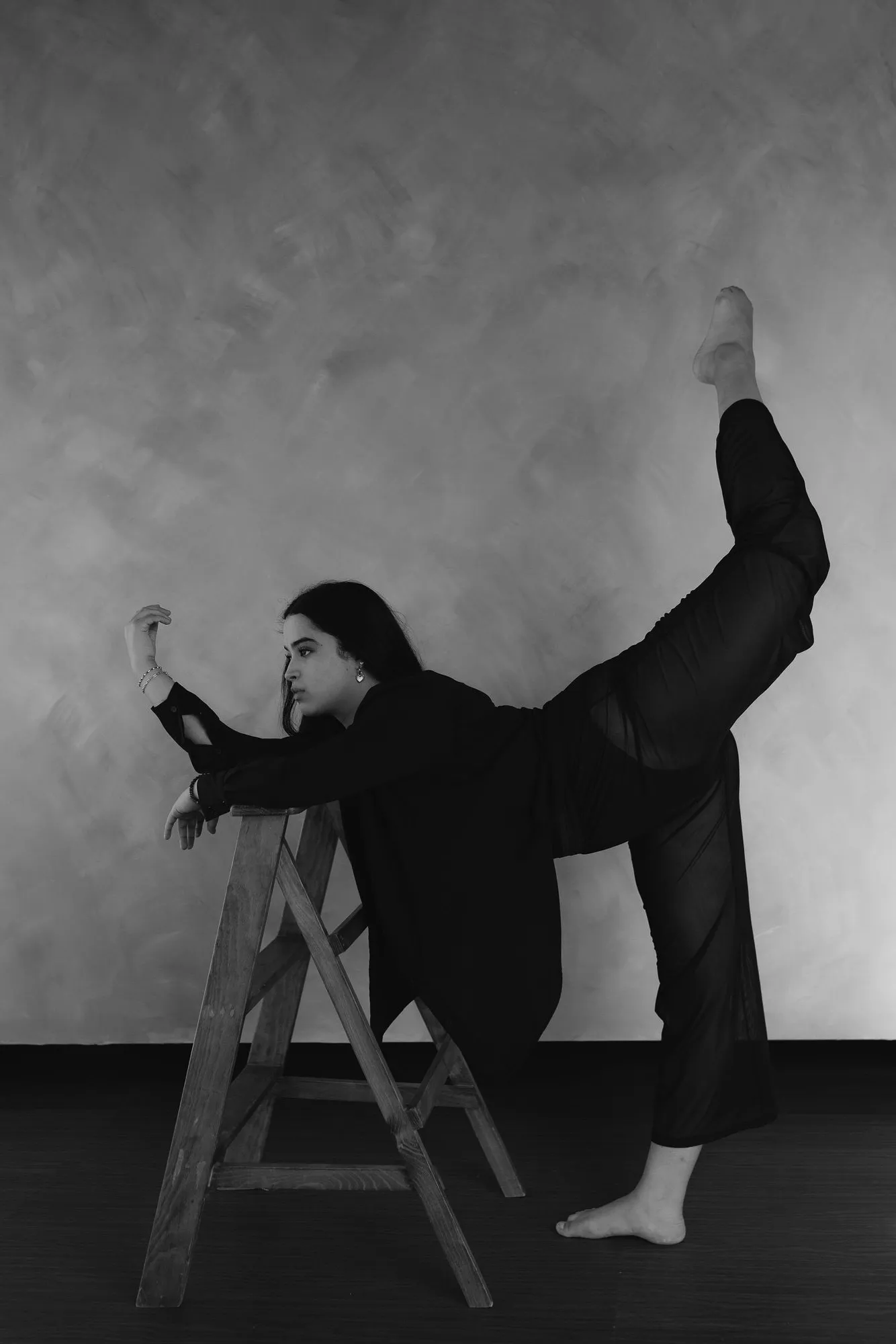 En blanco y negro, una mujer hace un arabesco apoyada en una escalera de madera, frente a una pared texturizada.
