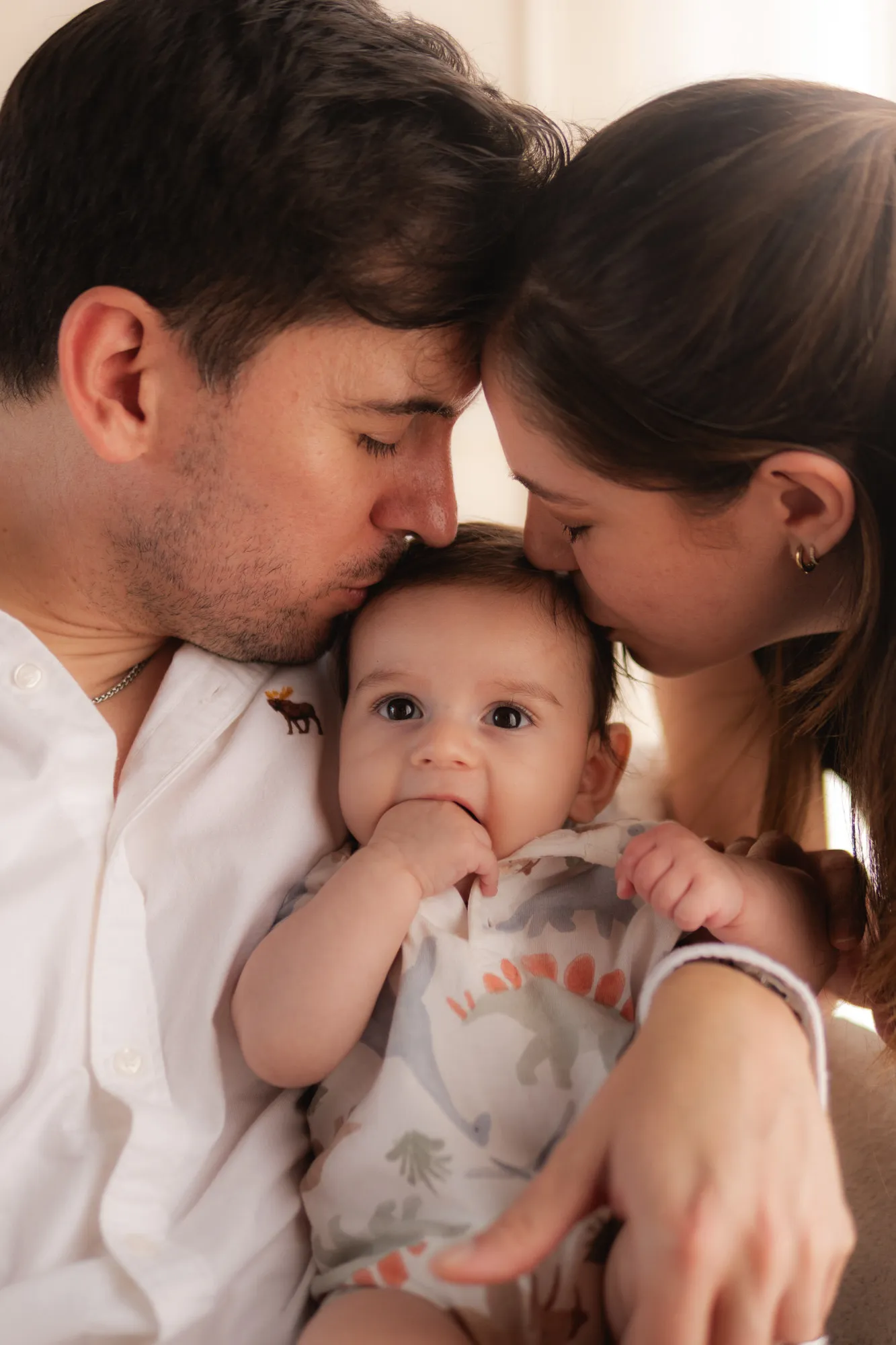 Un papá y una mamá besan la cabeza de su bebé, que mira a la cámara.