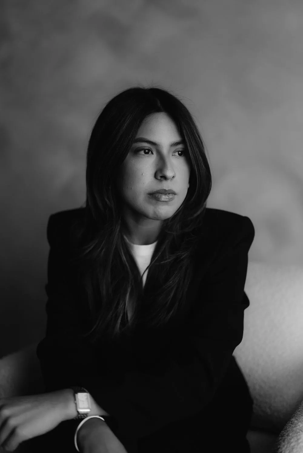 En blanco y negro, una mujer con saco oscuro y cabello largo mira de frente, sentada.