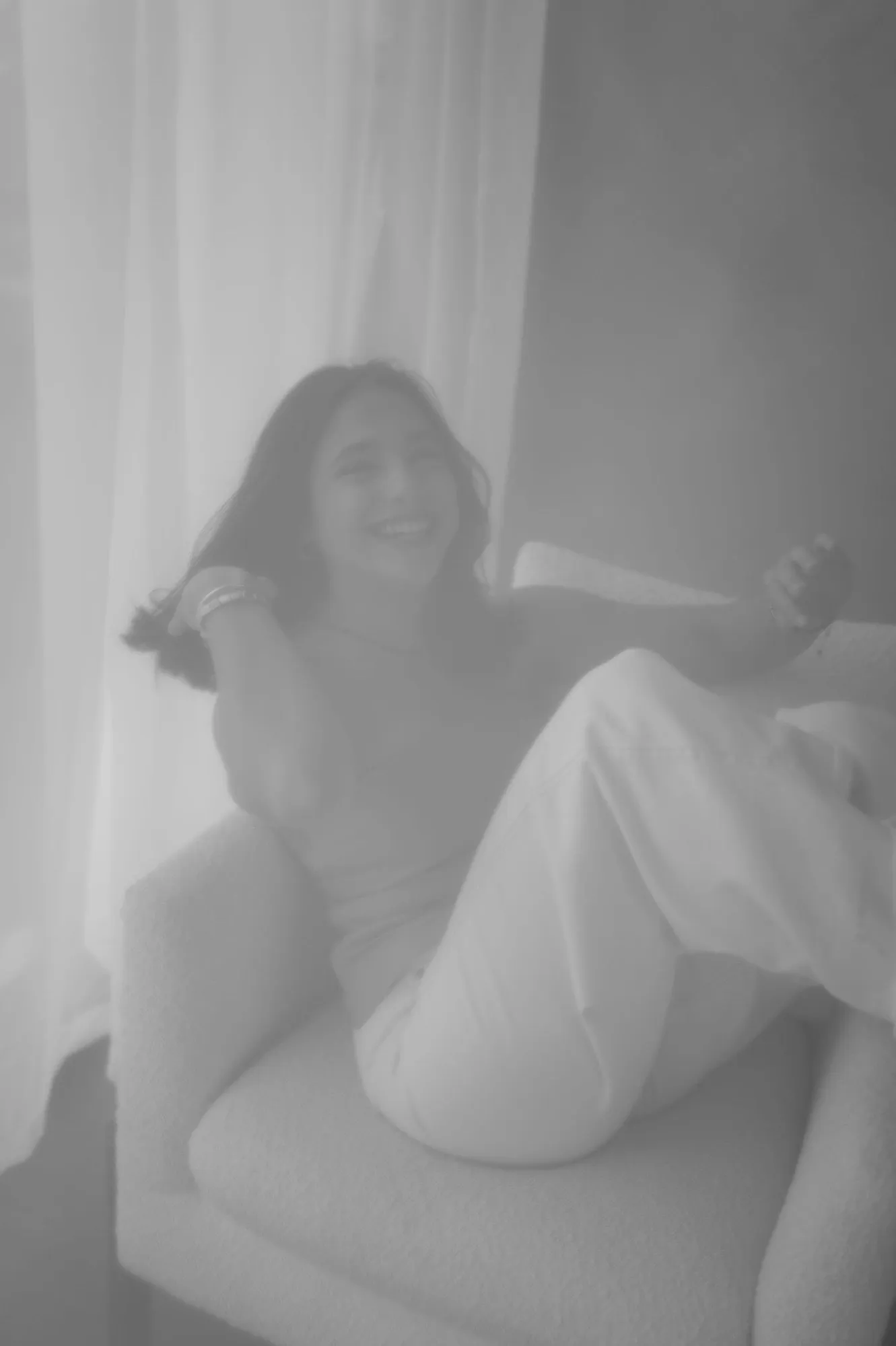 En blanco y negro, una mujer sentada en un sillón junto a una cortina, con la imagen suave y desvanecida.