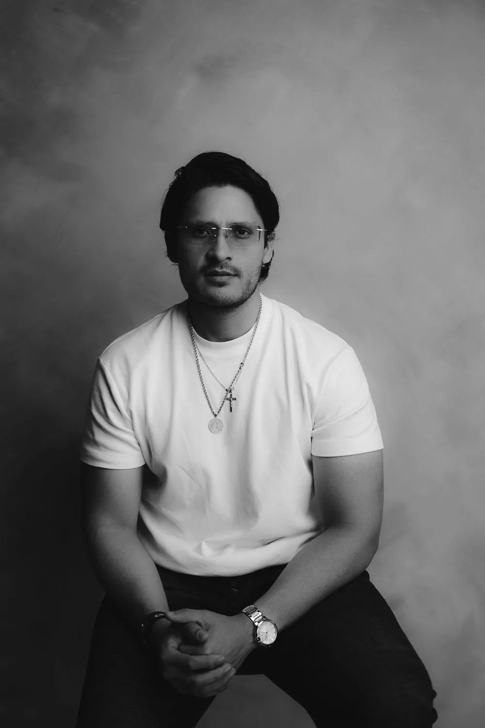 En blanco y negro, un hombre con playera blanca y cadena, sentado con las manos juntas, mira a la cámara.