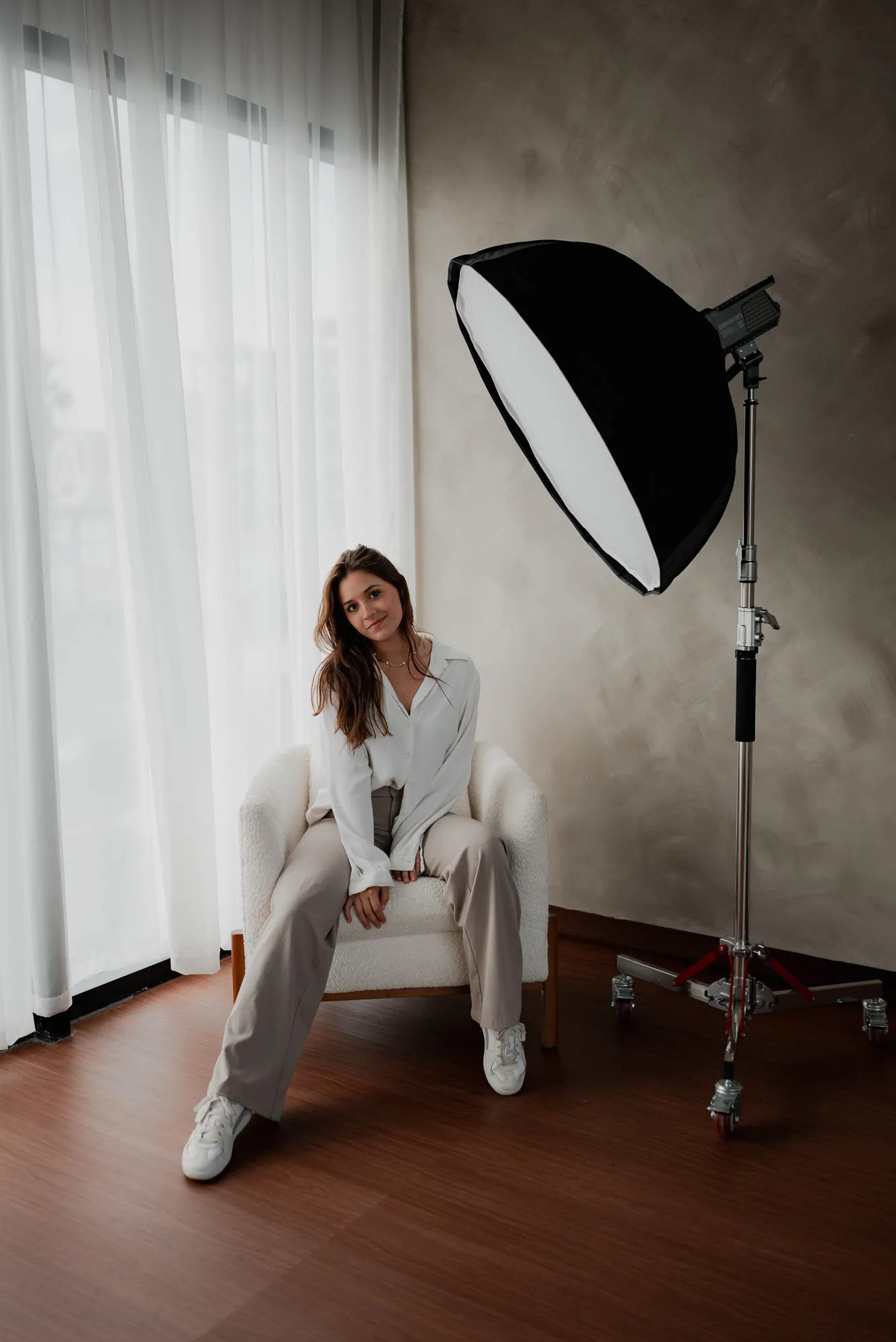 Una mujer sentada en un sillón junto a una caja de luz y ventanales con cortinas.