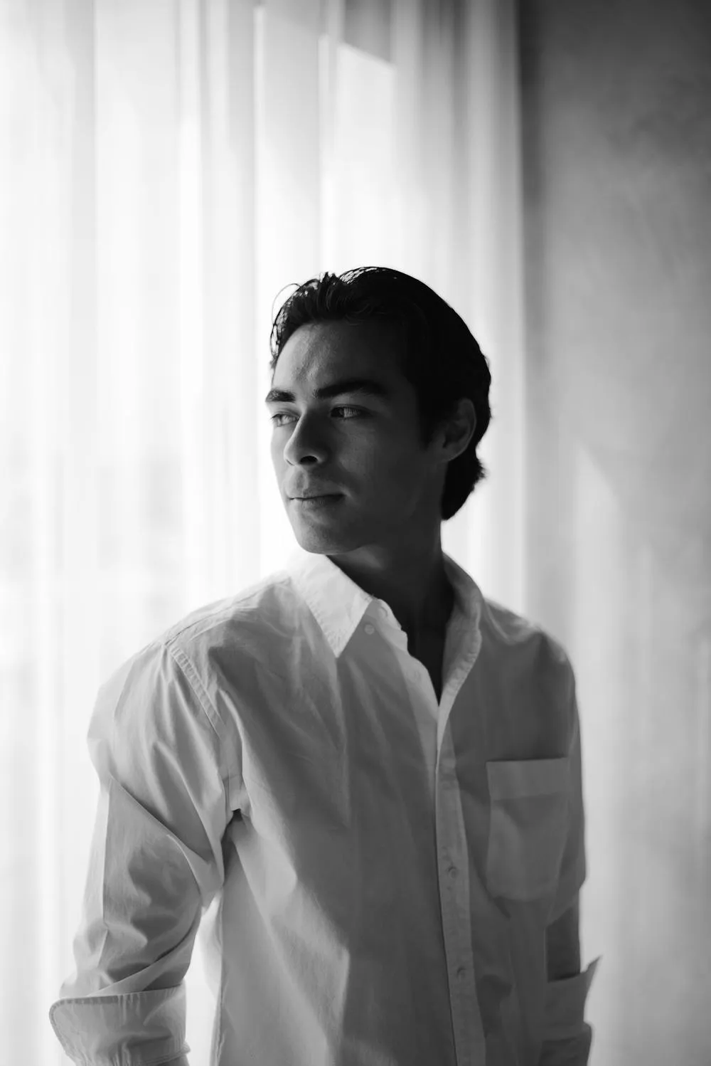 En blanco y negro, un hombre con camisa blanca mira hacia un lado, frente a una cortina.