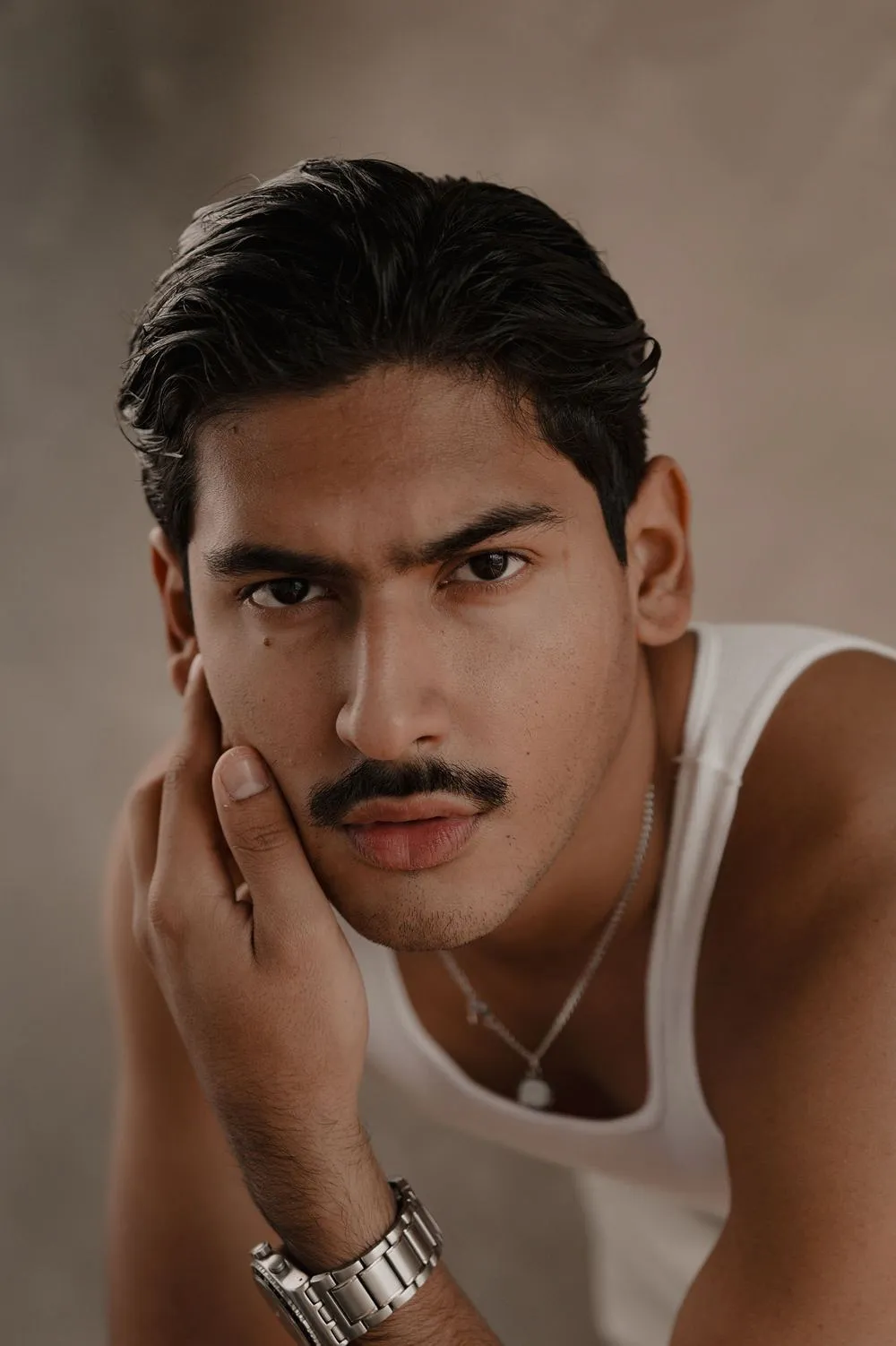 Primer plano de un hombre con bigote y playera blanca que apoya la cara en su mano y mira a la cámara.