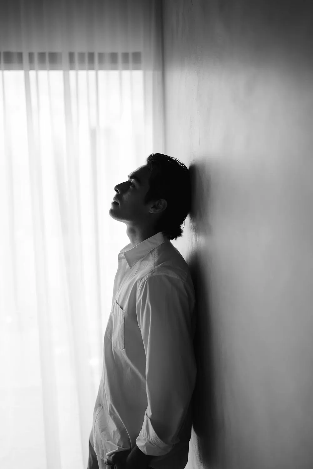 En blanco y negro, un hombre recargado en la pared, de perfil, mira hacia arriba.