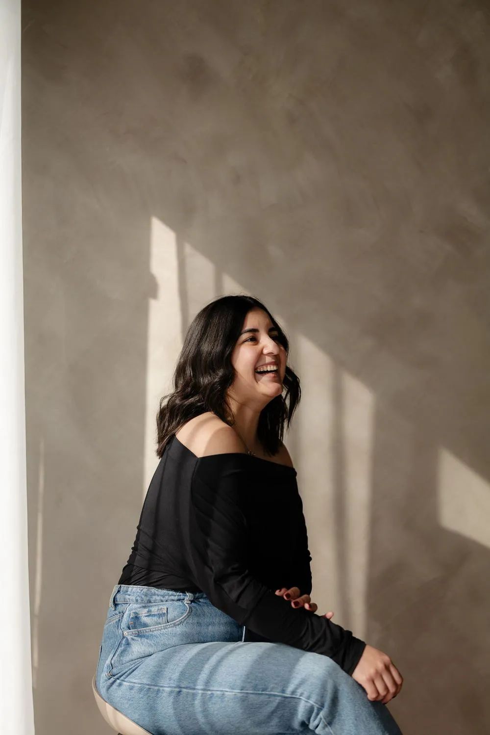 Una mujer ríe sentada de lado, con blusa negra y jeans, frente a una pared con luz y sombras.
