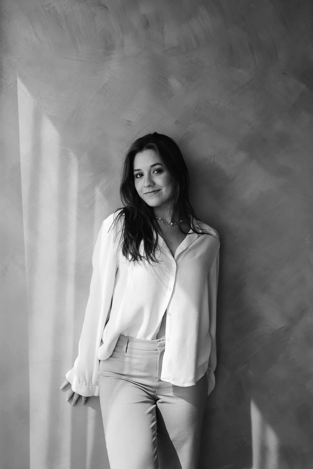 En blanco y negro, una mujer con blusa blanca y pantalón claro, de pie y recargada en una pared.