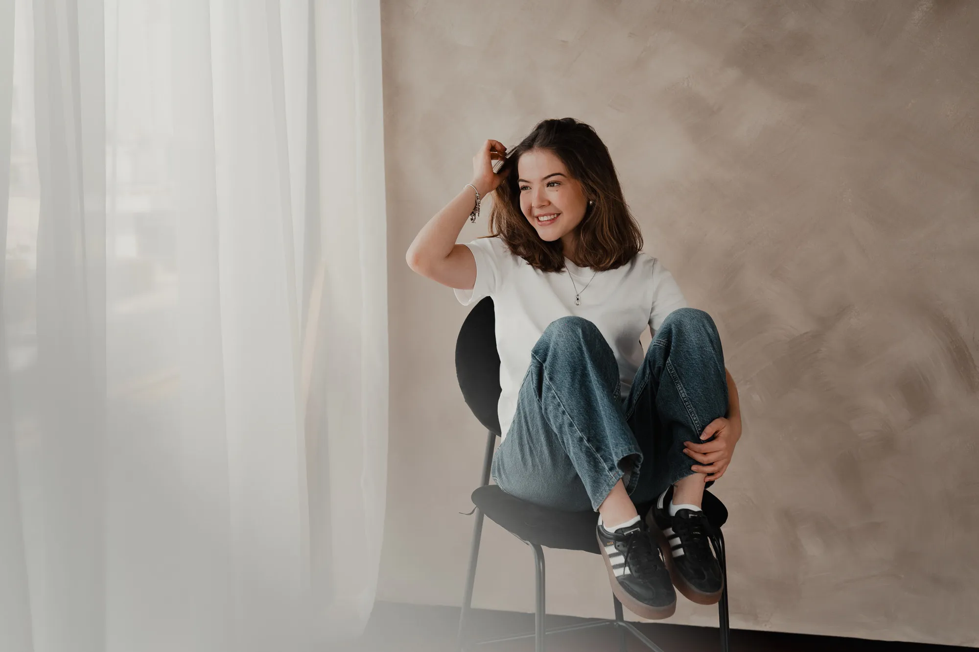 Una mujer con jeans y playera blanca, sentada en una silla negra, ríe frente a una pared color arena.