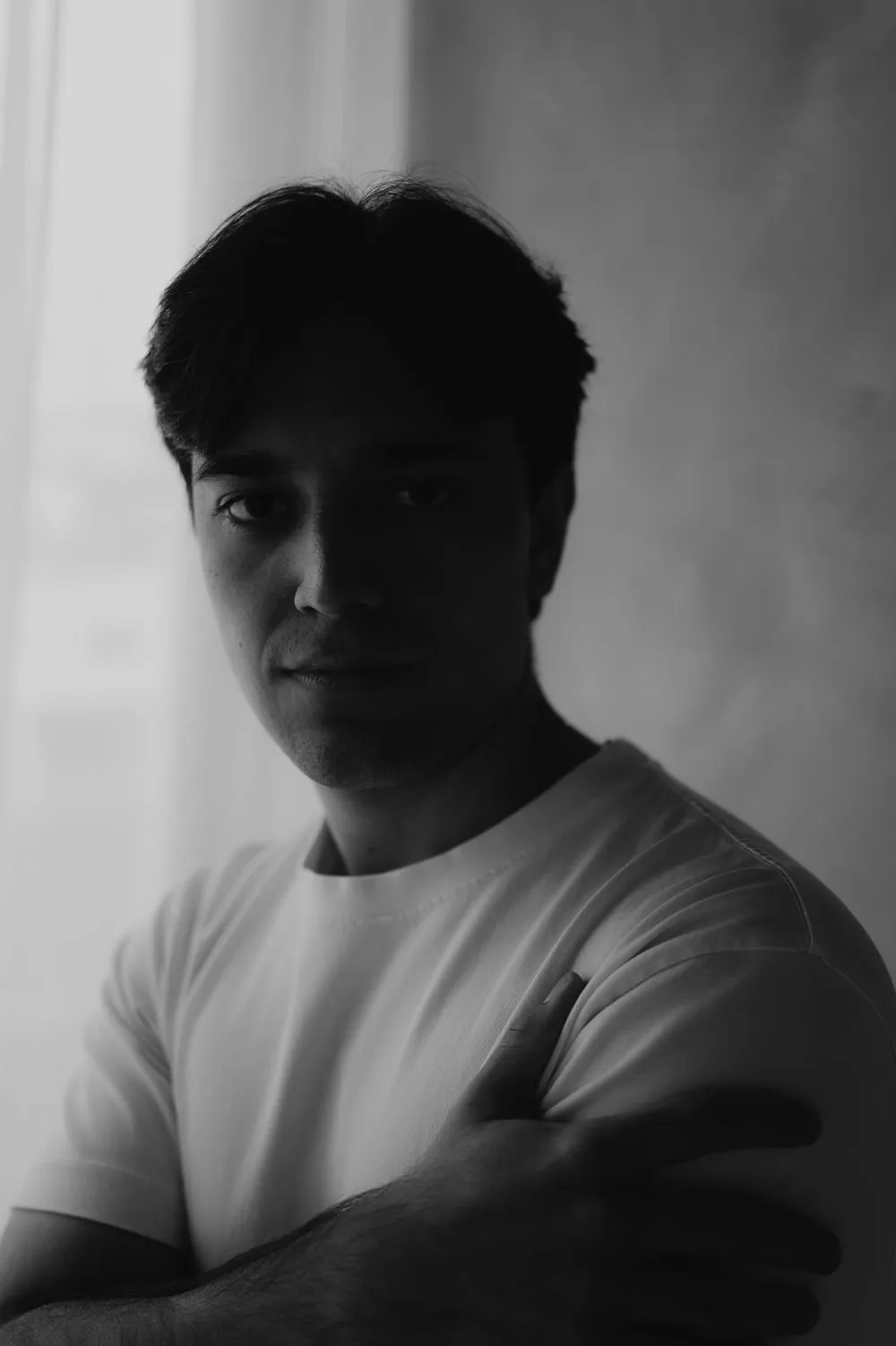 En blanco y negro, un joven con playera blanca y los brazos cruzados mira a la cámara.
