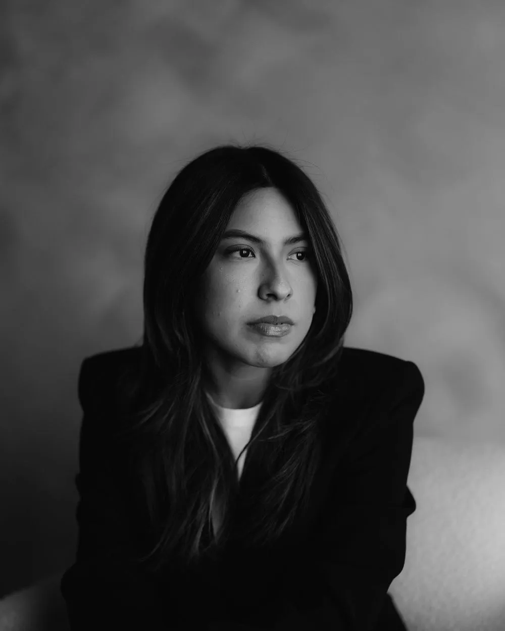 En blanco y negro, una mujer con saco oscuro y cabello largo mira de frente, sentada.